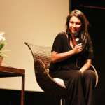 Foto de uma mulher palestrante sentada com um microfone á mão.