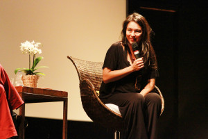 Foto de uma mulher palestrante sentada com um microfone á mão.