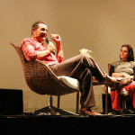 Foto onde á ao centro um palestrante homem, sentado com um microfone á mão e uma mulher á direita em um palco.