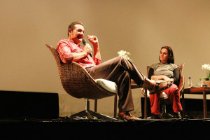 Foto onde á ao centro um palestrante homem, sentado com um microfone á mão e uma mulher á direita em um palco.