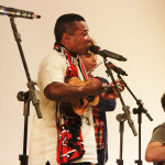 Foto de uma banda tocando , ao centro á um homem negro tocando cavaquinho uma mulher ao lado tocando flauta e á direita um homem sentado tocando violão .