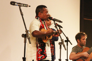 Foto de uma banda tocando , ao centro á um homem negro tocando cavaquinho uma mulher ao lado tocando flauta e á direita um homem sentado tocando violão .