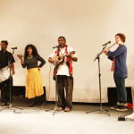 Foto de uma banda tocando em um palco , composta de cinco membros, á esquerda há um homem tocando percussão e uma mulher com um chocalho , ao centro há um homem tocando e um cavaquinho e cantando e á direita á uma mulher tocando uma flauta e um homem tocando violão sentado em um banco.