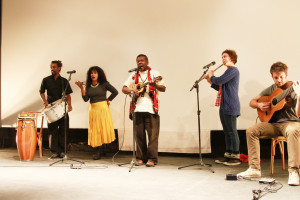 Foto de uma banda tocando em um palco , composta de cinco membros, á esquerda há um homem tocando percussão e uma mulher com um chocalho , ao centro há um homem tocando e um cavaquinho e cantando e á direita á uma mulher tocando uma flauta e um homem tocando violão sentado em um banco.