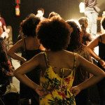 Foto mostrando um grupo de mulheres de costas com as mãos á cintura observando uma banda a tocar no palco.