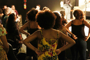 Foto mostrando um grupo de mulheres de costas com as mãos á cintura observando uma banda a tocar no palco.