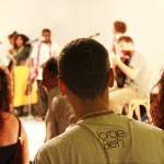 Foto onde há um grupo de pessoas assistindo uma banda ao fundo tocar.