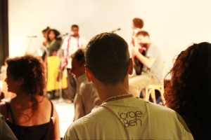 Foto onde há um grupo de pessoas assistindo uma banda ao fundo tocar.