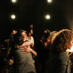 Foto mostrando inúmeras pessoas se interagindo , onde há abraços e comprimentos ao centro da imagem entre duas mulheres.