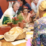 Foto composta por várias pessoas em volta da mesa fazendo batendo massa de pão, há vários pães prontos em cima da mesa.