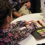 Foto de uma criança brincando com um estojo de lápis de cor que estão encima de uma mesa.