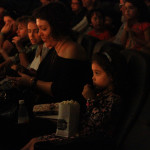 Foto com inúmeras crianças em uma sala de cinema , á esquerda da foto á uma menina com um saco de pipocas no colo .