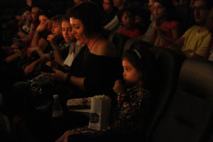 Foto com inúmeras crianças em uma sala de cinema , á esquerda da foto á uma menina com um saco de pipocas no colo .