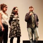 Foto composta de três pessoas , duas mulheres e um homem onde á direita da foto o homem está com um microfone á mão.