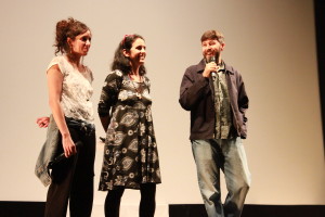 Foto composta de três pessoas , duas mulheres e um homem onde á direita da foto o homem está com um microfone á mão.