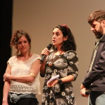 Foto composta de três pessoas , duas mulheres e um homem onde á mulher no centro da foto está com um microfone á mão.