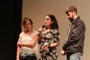 Foto composta de três pessoas , duas mulheres e um homem onde á mulher no centro da foto está com um microfone á mão.