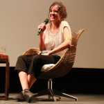 Foto de uma mulher sentada sorrindo com um microfone na mão.