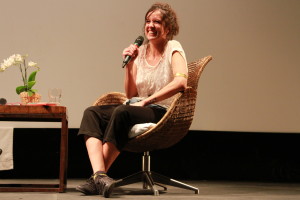 Foto de uma mulher sentada sorrindo com um microfone na mão.