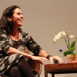 Foto de uma mulher sentada sorrindo com um microfone na mão.