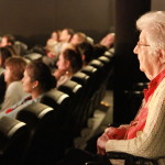 Foto de uma sala de cinema, onde á esquerda á uma senhora em pé observando a sessão.