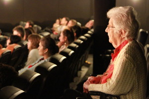 Foto de uma sala de cinema, onde á esquerda á uma senhora em pé observando a sessão.