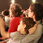 Foto de uma sala de cinema com muitas pessoas , o destaque da foto é uma mulher com seu filho no colo.