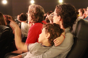 Foto de uma sala de cinema com muitas pessoas , o destaque da foto é uma mulher com seu filho no colo.