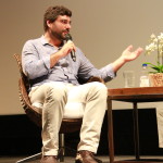 Foto ilustrando uma palestra , onde á um homem sentado em uma cadeira em um palco com um microfone á mão.