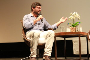 Foto ilustrando uma palestra , onde á um homem sentado em uma cadeira em um palco com um microfone á mão.