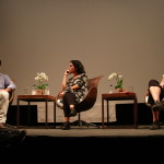 Foto ilustrando um palco onde á uma palestra , á esquerda um homem com um microfone na mão, uma mulher ao centro olhando para o homem da esquerda e á direita á outra mulher também observando o mesmo homem falando ao microfone.