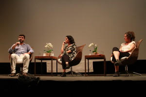 Foto ilustrando um palco onde á uma palestra , á esquerda um homem com um microfone na mão, uma mulher ao centro olhando para o homem da esquerda e á direita á outra mulher também observando o mesmo homem falando ao microfone.