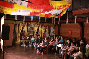 Foto mostrando pessoas assistindo algo em um telão.