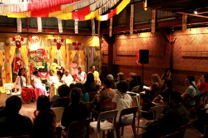 Foto ilustrando  o público total de uma palestra ,onde ao fundo há um palco composto de quatro mulheres e dois homens falando a um número de pessoas sentadas assistindo ao evento.