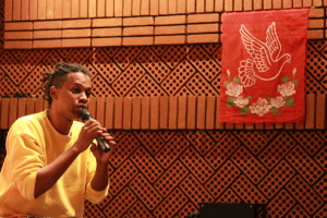 Foto onde há um homem negro com um microfone na mão falando ao público.