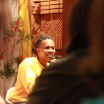 Foto ilustrando um diálogo entre pessoas , onde ao centro da imagem há um homem negro com um microfone na mão.