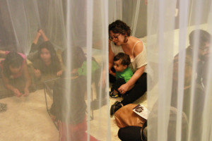 Foto com quatro crianças se interagindo , onde ao centro á uma mulher com um menino sentado em seu colo.