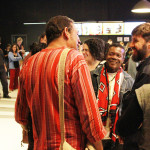 Foto ilustrando um diálogo entre pessoas , onde á um homem de costas á esquerda , um homem negro ao centro e outro homem á esquerda.