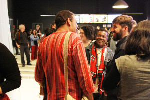 Foto ilustrando um diálogo entre pessoas , onde á um homem de costas á esquerda , um homem negro ao centro e outro homem á esquerda.