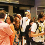 Foto mostrando um local onde várias pessoas estão se interagindo , onde ao centro há um homem e uma mulher tirando uma foto e á direita á um rapaz utilizando seu smartphone.