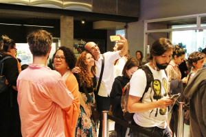 Foto mostrando um local onde várias pessoas estão se interagindo , onde ao centro há um homem e uma mulher tirando uma foto  e á direita  á um rapaz utilizando seu smartphone.