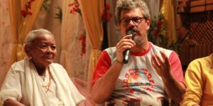 Uma senhora negra ao lado de um homem que fala ao microfone.