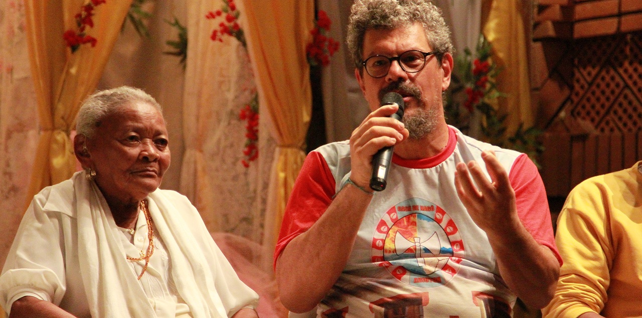 Uma senhora negra ao lado de um homem que fala ao microfone.