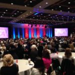 Foto de um grande salão de eventos, várias pessoas estão sentadas em volta de mesas redondas.