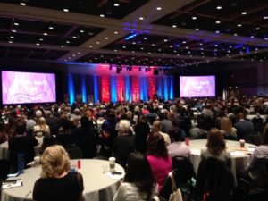 Foto de um grande salão de eventos, várias pessoas estão sentadas em volta de mesas redondas.