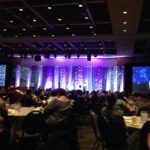 Foto de um grande salão de eventos com palco iluminado em lilás.