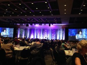 Foto de um grande salão de eventos com palco iluminado em lilás.