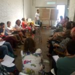 Foto de um homem em frente de dois quadros, fala para uma turma de pessoas sentadas que o observar.