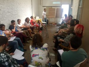 Foto de um homem em frente de dois quadros, fala para uma turma de pessoas sentadas que o observar.