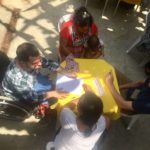 Foto composta por quatro pessoas sentada em uma mesa, são dois homens e duas mulheres o homem da esquerda esta sentado em uma cadeira de rodas a sua esquerda uma mulher segura um bebê, ambos estão discutindo.